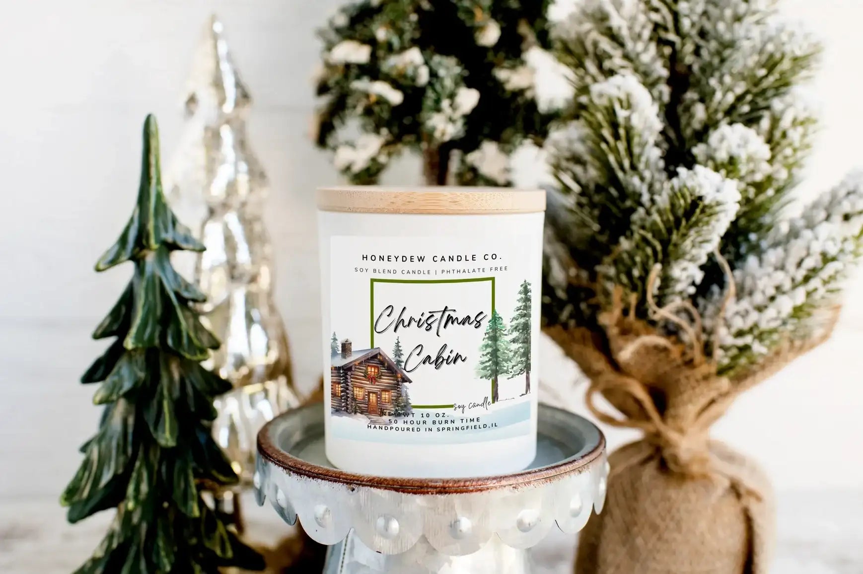 Honeydew Candle Co Christmas Cabin candle on a small white stand with decorative trees in the background.