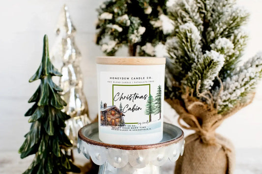 Honeydew Candle Co Christmas Cabin candle on a small white stand with decorative trees in the background.