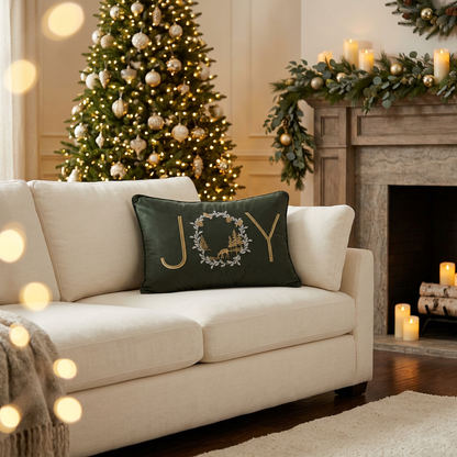 Olive-green rectangular Christmas throw pillow with gold embroidered JOY lettering and a white evergreen wreath featuring a deer, gold forest trees, pinecones, and birds.
