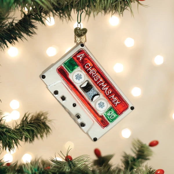 A Christmas cassette tape ornament hanging from a tree branch, featuring a white cassette body with red, green, and silver details and the text 'A CHRISTMAS MIX 90'.