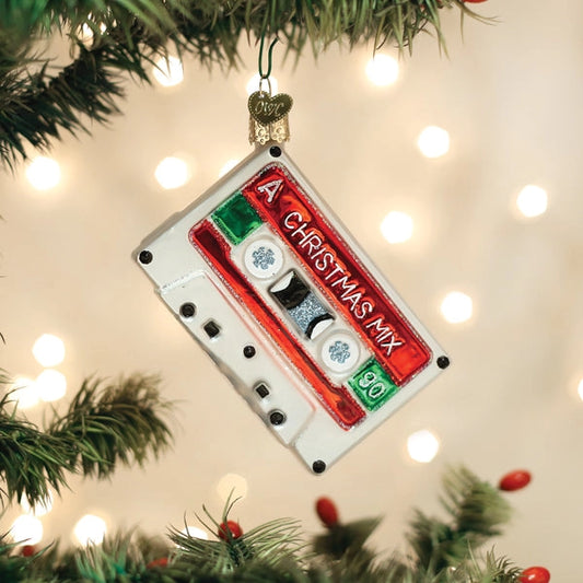 A Christmas cassette tape ornament hanging from a tree branch, featuring a white cassette body with red, green, and silver details and the text 'A CHRISTMAS MIX 90'.