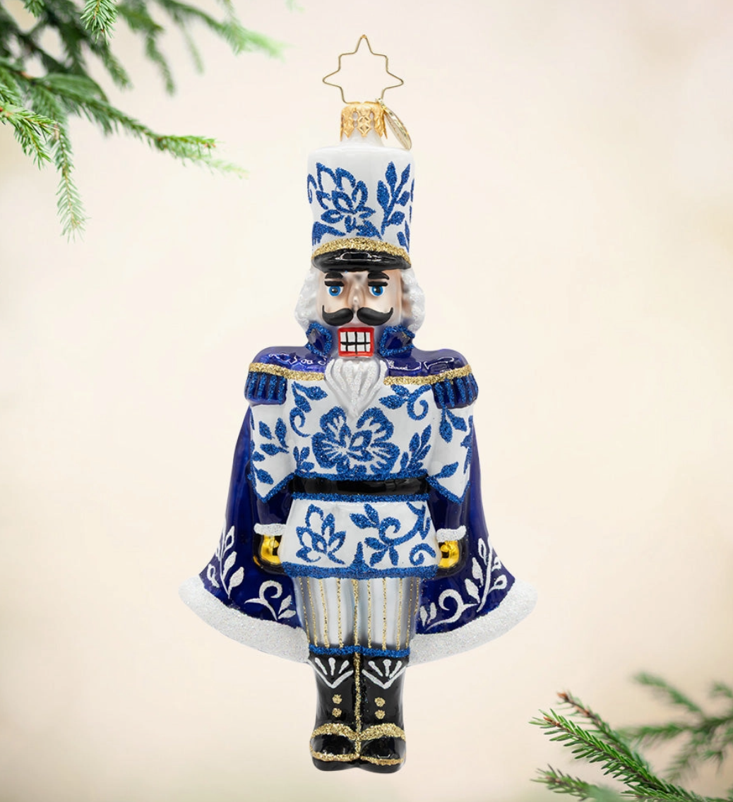 A ceramic nutcracker Christmas ornament with a blue and white floral pattern, gold trim, and a gold star topper, shown hanging against a soft beige background with pine branches at the edges.