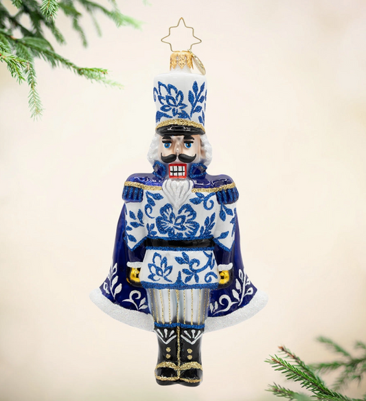 A ceramic nutcracker Christmas ornament with a blue and white floral pattern, gold trim, and a gold star topper, shown hanging against a soft beige background with pine branches at the edges.