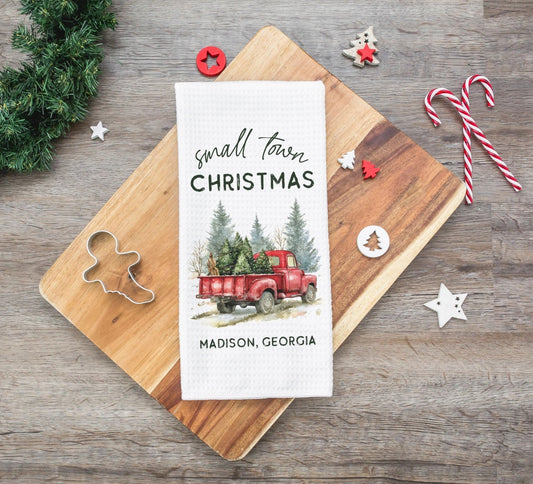 A white kitchen tea towel with a 'small town CHRISTMAS' graphic featuring a red truck and pine trees, laid flat on a wooden cutting board. The board is styled on a rustic gray wood table with holiday decorations including a green wreath, candy canes, and wooden ornaments.
