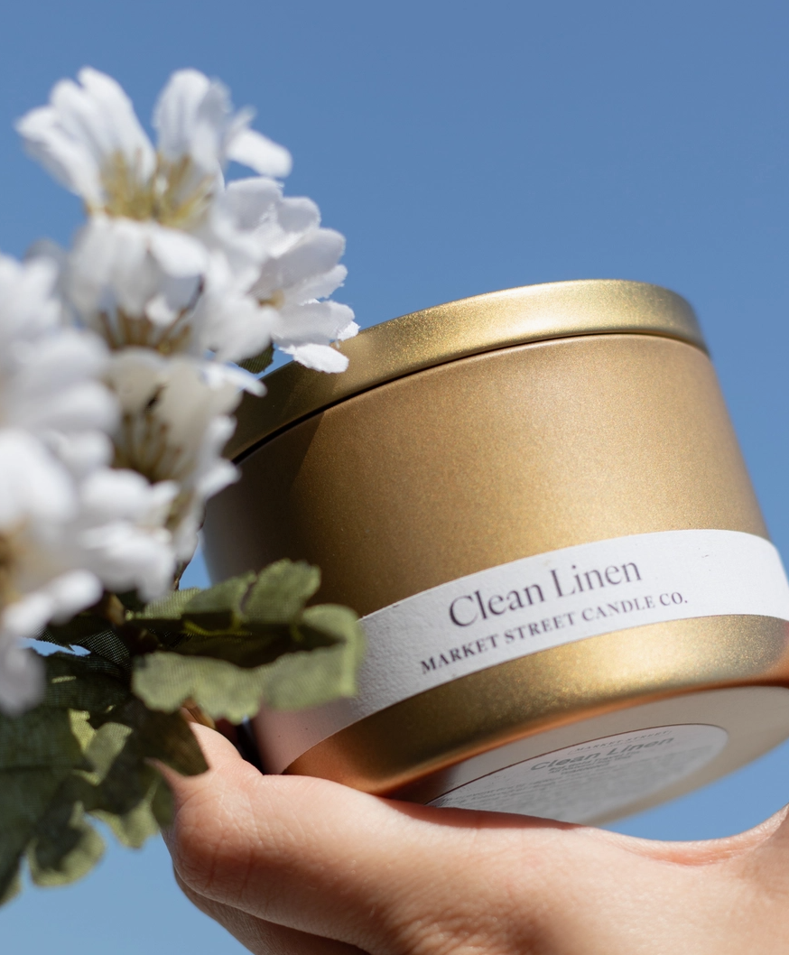 Market Street Candle Co. Clean Linen Travel Tin soy candle in a gold metal tin surrounded by green foliage and small white flowers.