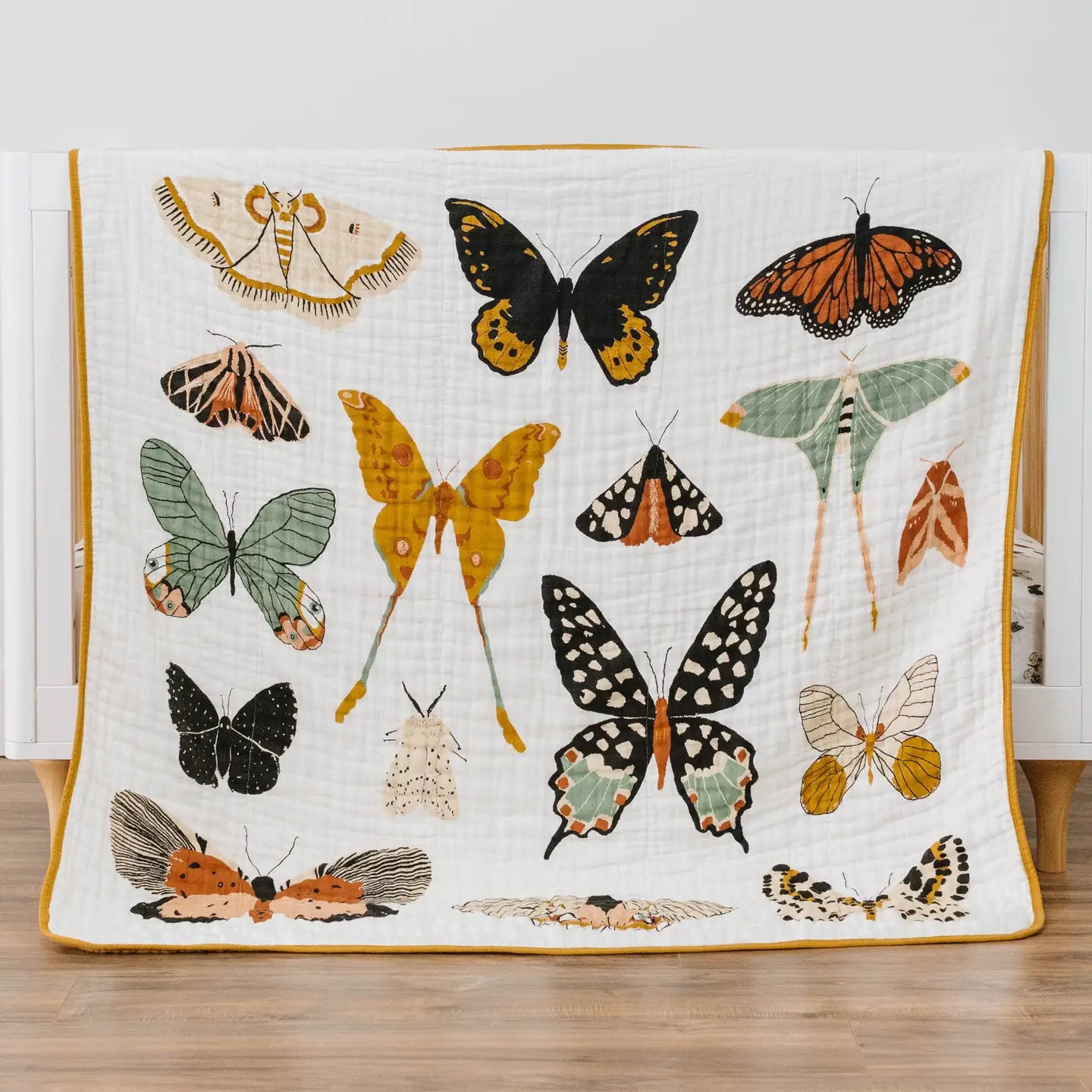 A white muslin quilt with a yellow border, decorated with various colorful butterflies and moths, displayed on a white crib in a nursery setting.
