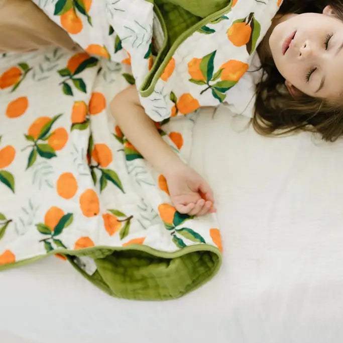 A child lying on a white bed wrapped in a quilted blanket featuring an orange and green citrus print.