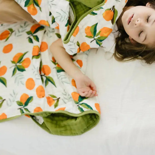 A child lying on a white bed wrapped in a quilted blanket featuring an orange and green citrus print.