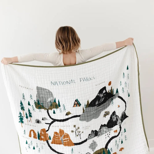A woman holding up a large white muslin blanket with a printed National Parks map design, shown from behind against a white wall.