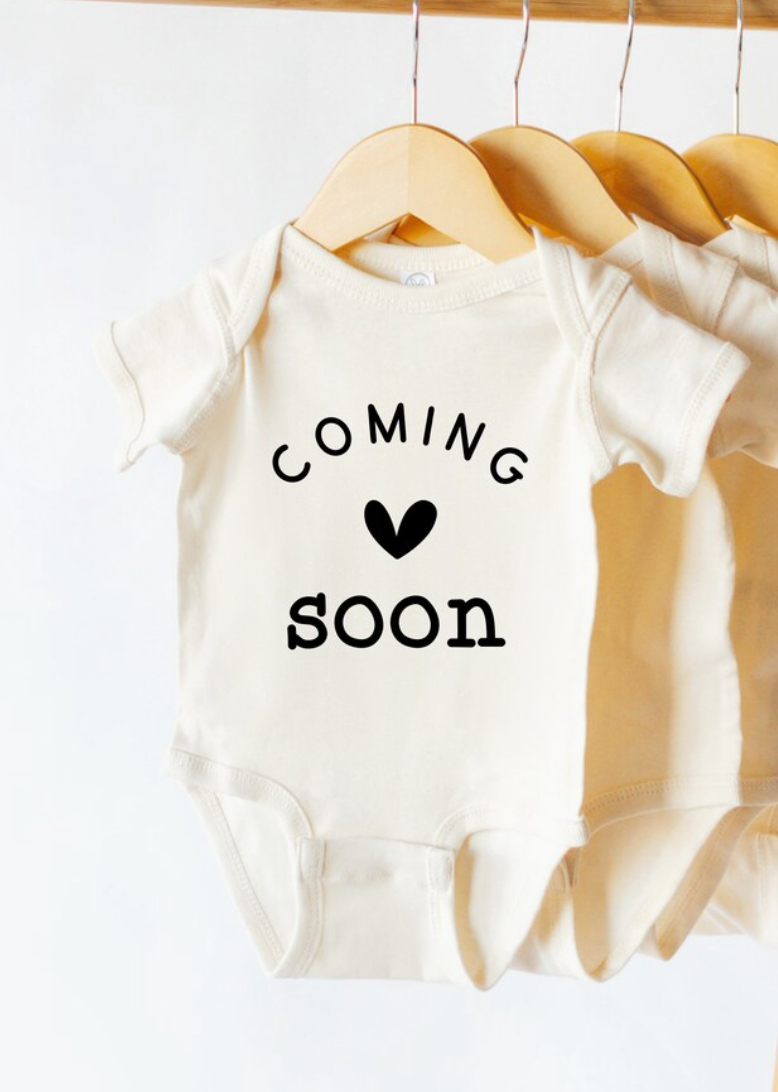 Baby onesie with 'Coming soon' text on a hanger against a white background
