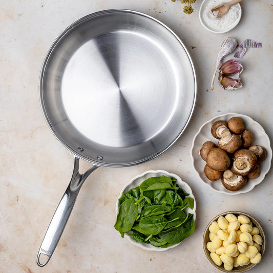 Stainless steel frying pan on a light surface with bowls of spinach, mushrooms, and gnocchi.