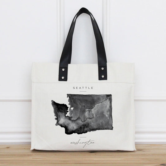 Tote bag with a map of Washington state and 'Seattle' printed on it, against a white wooden panel background.