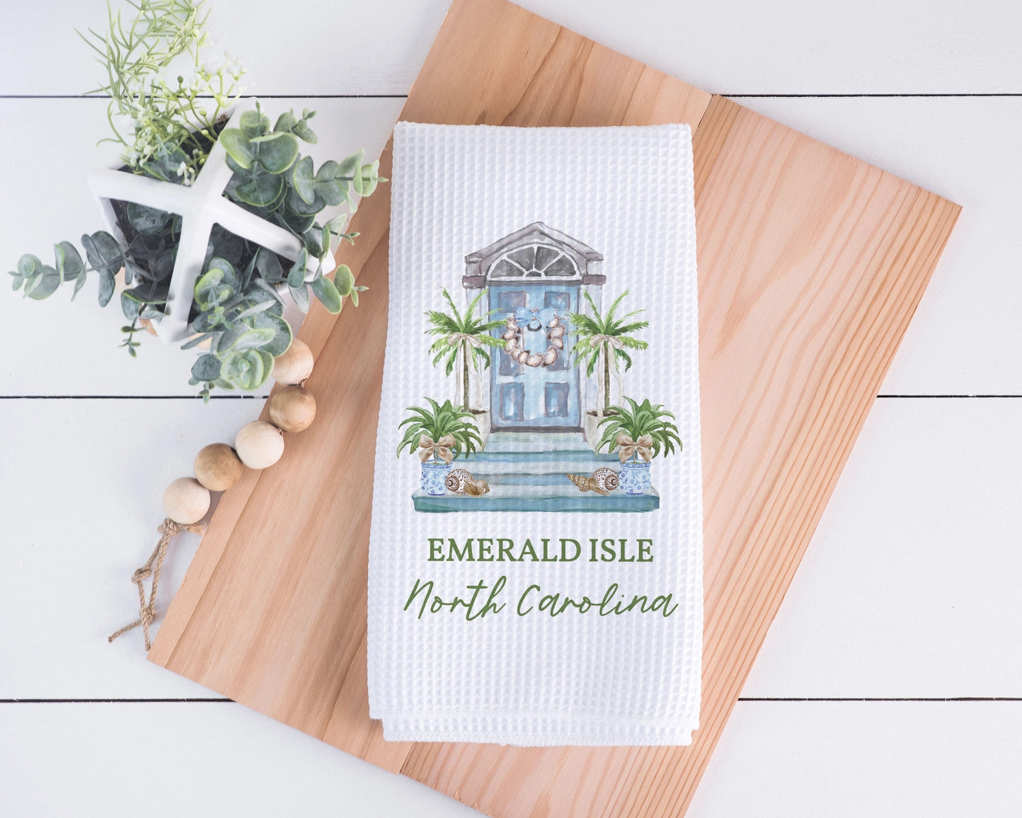 A white waffle-weave kitchen towel featuring a printed illustration of a blue front door with palm trees and the text 'EMERALD ISLE North Carolina,' styled on a wooden board with decorative greenery and beads.