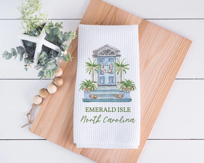 A white waffle-weave kitchen towel featuring a printed illustration of a blue front door with palm trees and the text 'EMERALD ISLE North Carolina,' styled on a wooden board with decorative greenery and beads.