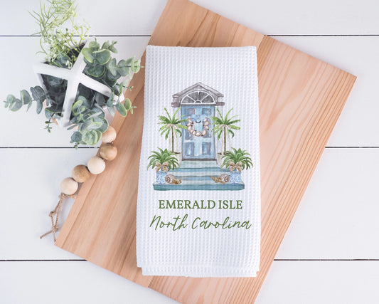 A white waffle-weave kitchen towel featuring a printed illustration of a blue front door with palm trees and the text 'EMERALD ISLE North Carolina,' styled on a wooden board with decorative greenery and beads.