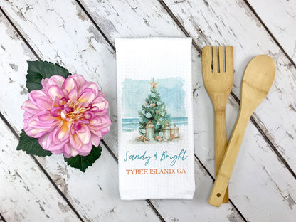 A white kitchen towel with a watercolor Christmas tree design and the text 'Sandy & Bright' and 'TYBEE ISLAND, GA' is displayed in a flat-lay arrangement on a white wood background, flanked by a pink flower and wooden utensils.