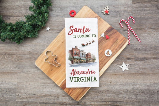 A personalized Christmas tea towel with a holiday illustration and the text 'Santa is coming to Alexandria Virginia' is displayed on a wooden cutting board. The cutting board is styled on a rustic wood table with Christmas decorations including a green pine wreath, candy canes, and wooden ornaments.