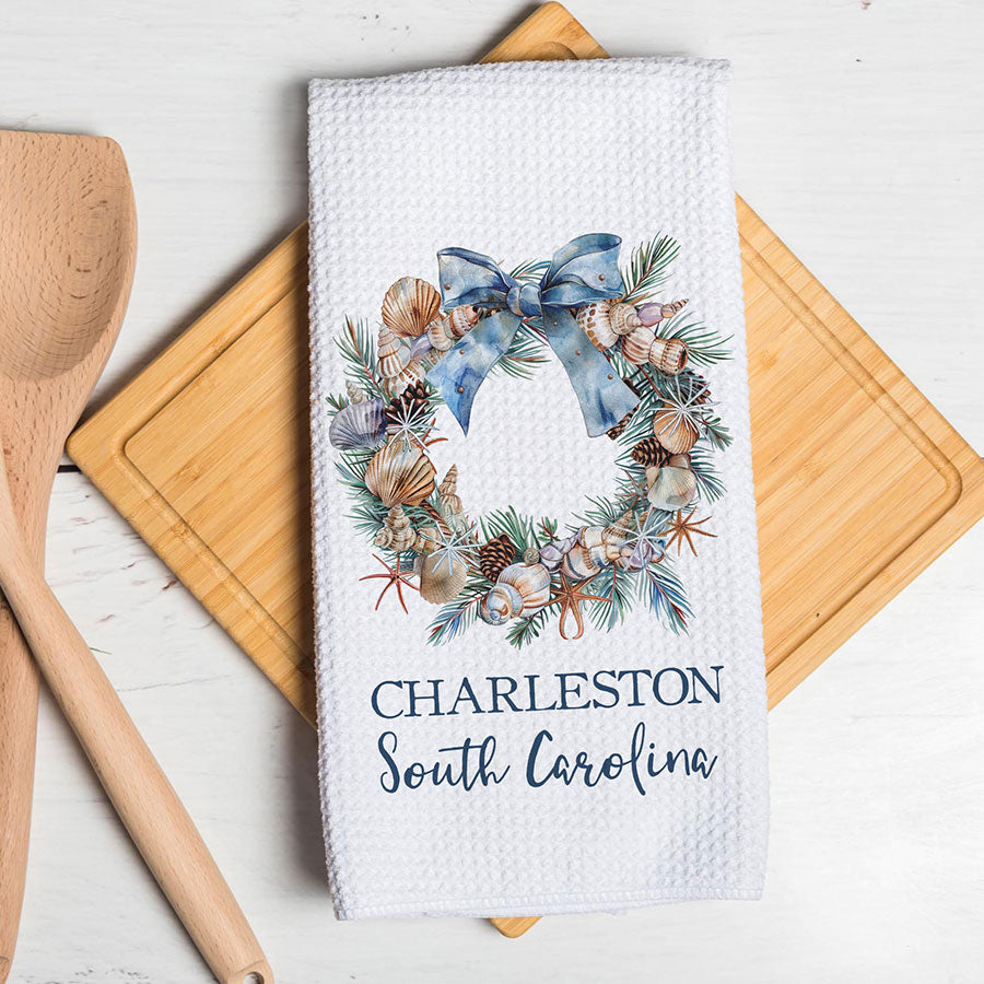 A white waffle-weave kitchen towel with a watercolor wreath design featuring seashells, pinecones, and a blue bow, with the text 'CHARLESTON South Carolina' printed below. The towel is styled on a wooden cutting board with a wooden spoon on a white wooden table.