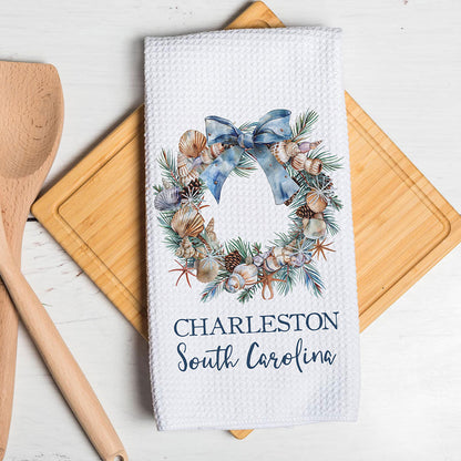 A white waffle-weave kitchen towel with a watercolor wreath design featuring seashells, pinecones, and a blue bow, with the text 'CHARLESTON South Carolina' printed below. The towel is styled on a wooden cutting board with a wooden spoon on a white wooden table.