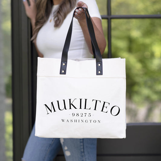 White tote bag with 'Mukilteo 98275 Washington' text held by a person outdoors.