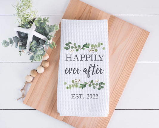 White towel with 'Happily ever after EST. 2022' text on a wooden board with decorative elements.