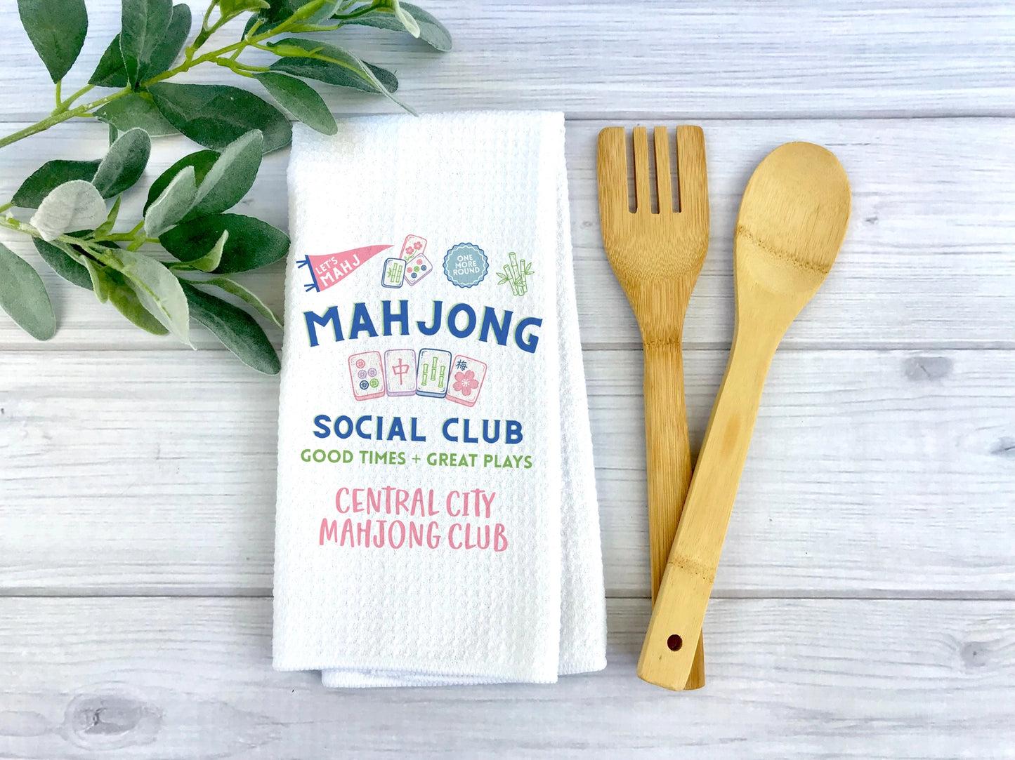 A white waffle-weave tea towel printed with colorful mahjong-themed text and graphics, styled on a light gray wood surface with green leaves on the left and a bamboo utensil set on the right.