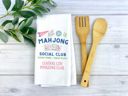 A white waffle-weave tea towel printed with colorful mahjong-themed text and graphics, styled on a light gray wood surface with green leaves on the left and a bamboo utensil set on the right.