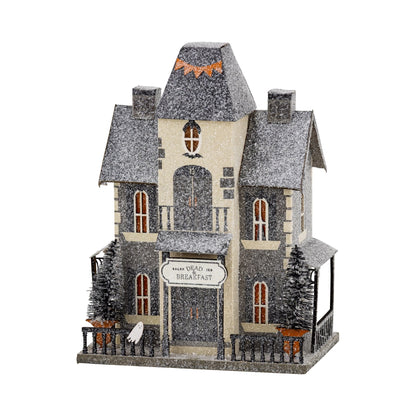 A decorative haunted house holiday ornament with a snow-covered gray and beige exterior, arched front door, small front porch, fence, miniature trees, and a sign reading 'Have You Been My Breakfast?'.