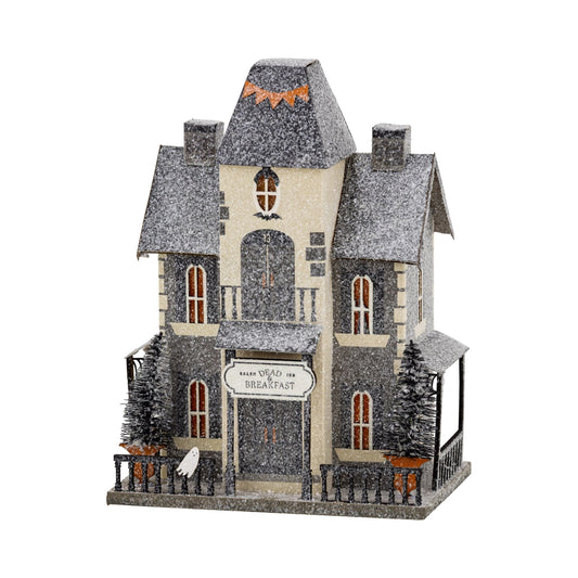 A decorative haunted house holiday ornament with a snow-covered gray and beige exterior, arched front door, small front porch, fence, miniature trees, and a sign reading 'Have You Been My Breakfast?'.