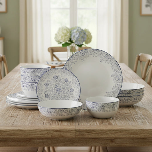 A coordinated dinnerware set arranged on a wooden dining table in a softly lit room. The set includes plates, bowls, and stacked pieces with a white base and delicate blue floral pattern around the rims and across the surfaces.