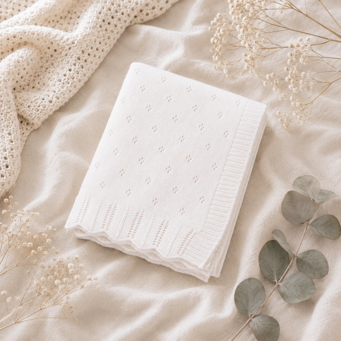 A folded white textured knit blanket with a delicate eyelet pattern and ribbed scalloped border, styled on a soft cream fabric with dried flowers and eucalyptus leaves.