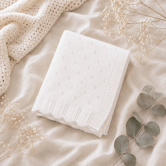 A folded white textured knit blanket with a delicate eyelet pattern and ribbed scalloped border, styled on a soft cream fabric with dried flowers and eucalyptus leaves.