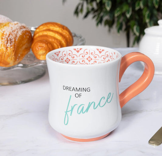 White mug with orange interior and handle, featuring 'Dreaming of France' text, on a table with pastries and a teapot.