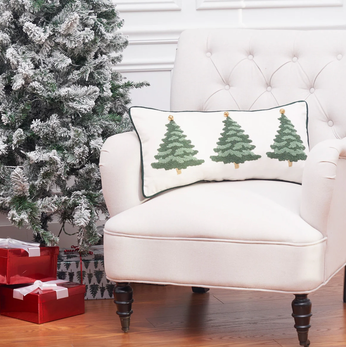 Christmas tree lumbar throw pillow displayed on an ivory accent chair beside a flocked Christmas tree in an elegant holiday living room.