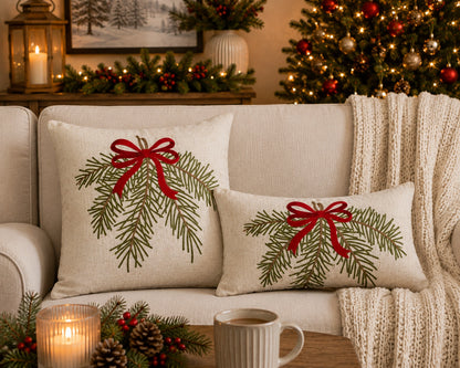 Two beige throw pillows with embroidered green pine branches and red ribbon bows, styled on a cream sofa in a cozy holiday living room setting with a lit mug, candles, and a Christmas tree in the background.