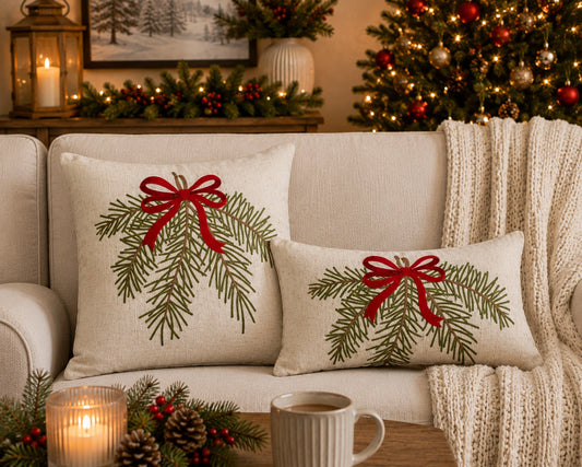 Two beige throw pillows with embroidered green pine branches and red ribbon bows, styled on a cream sofa in a cozy holiday living room setting with a lit mug, candles, and a Christmas tree in the background.