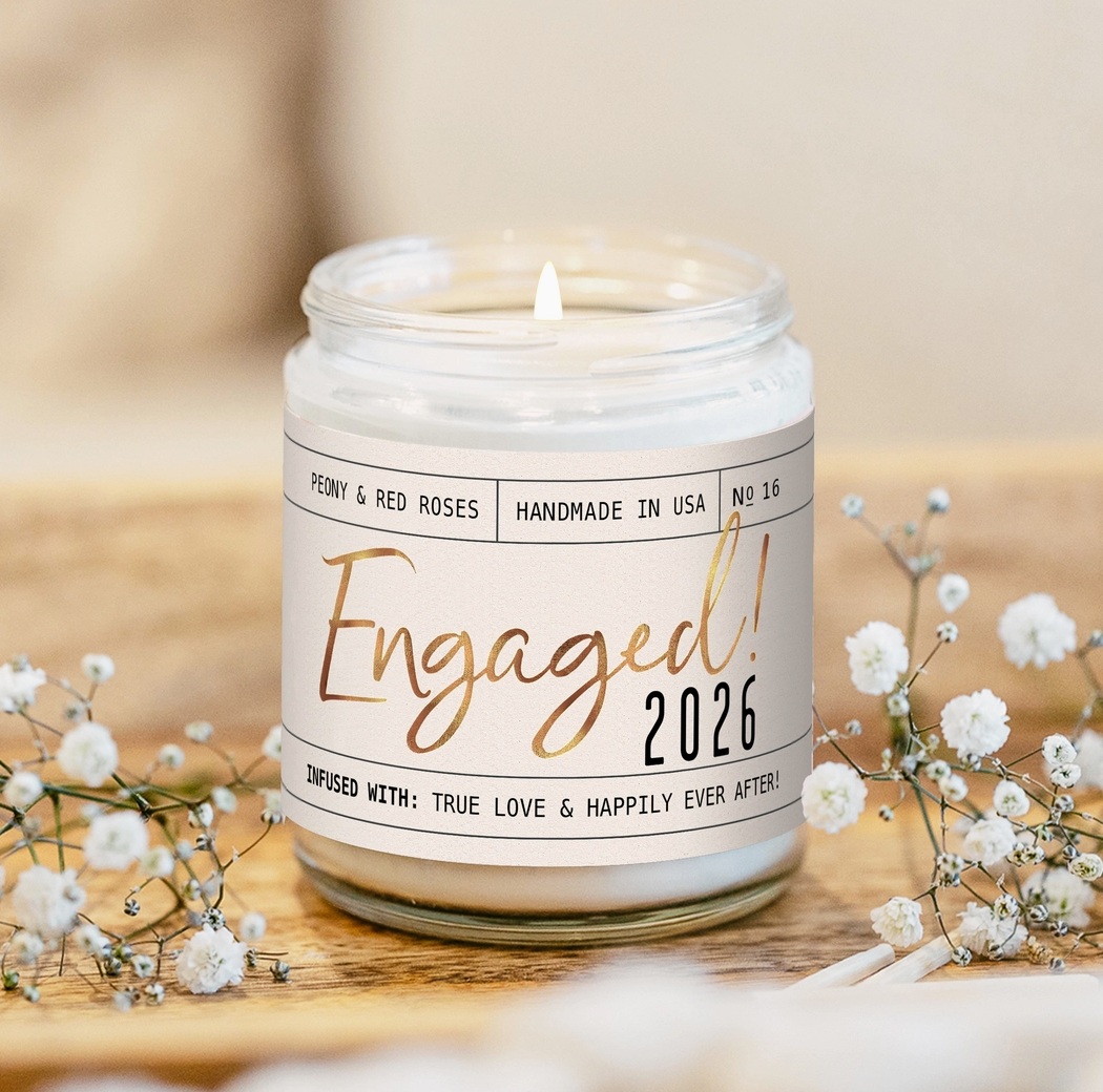 Candle in a jar labeled 'Engaged! 2026' with floral decorations on a wooden surface.