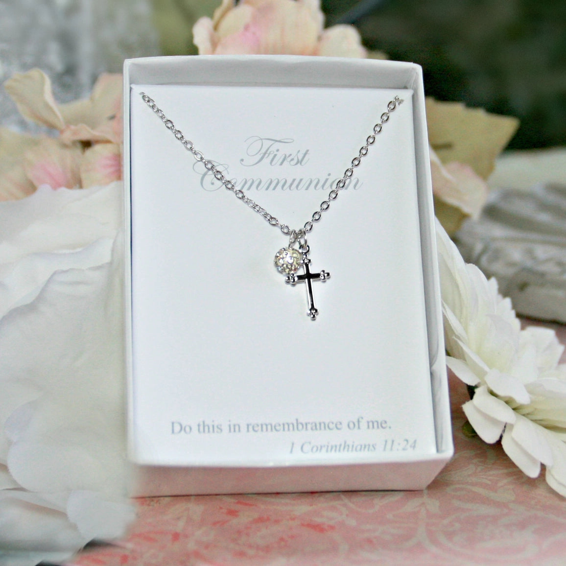 Necklace with cross pendant in a white box with text, surrounded by flowers.