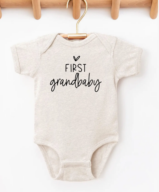 Beige baby onesie with 'First grandbaby' text on a white background