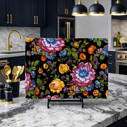 A rectangular floral art print or poster displayed on a small easel stand in a modern kitchen setting. The print features a dense, colorful floral pattern with pink, blue, yellow, and orange flowers on a black background.