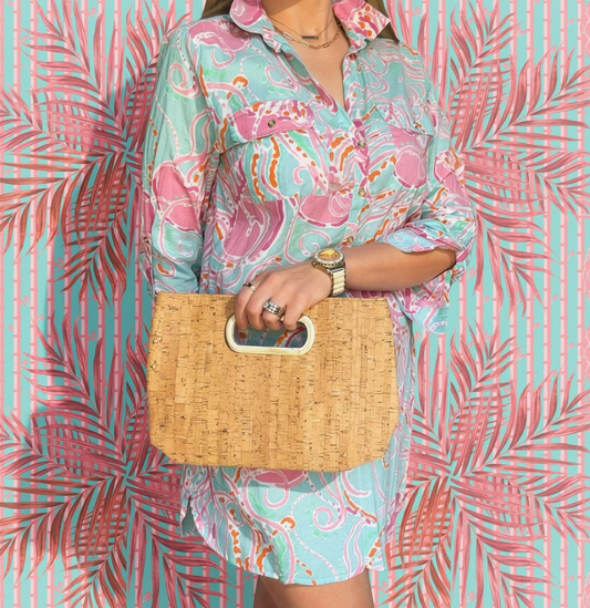 Person wearing a colorful patterned outfit holding a woven bag against a pink and green leafy background