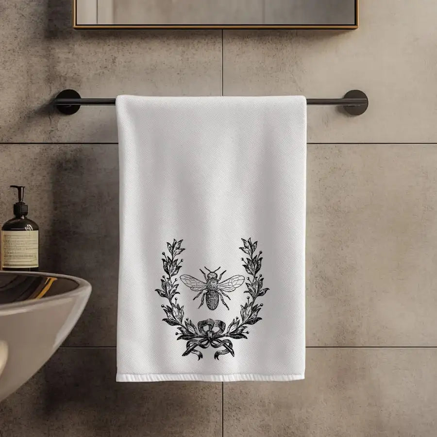 White towel with black laurel wreath and bee design hanging on a towel rack in a bathroom.