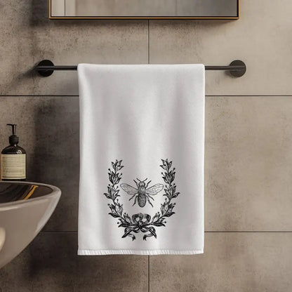 White towel with black laurel wreath and bee design hanging on a towel rack in a bathroom.