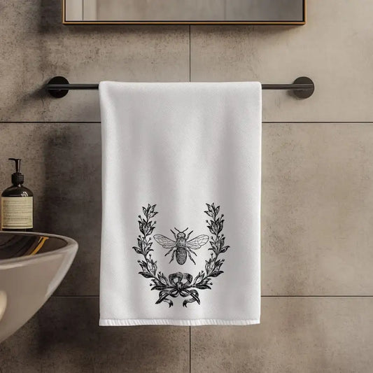 White towel with black laurel wreath and bee design hanging on a towel rack in a bathroom.