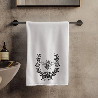 White towel with black laurel wreath and bee design hanging on a towel rack in a bathroom.