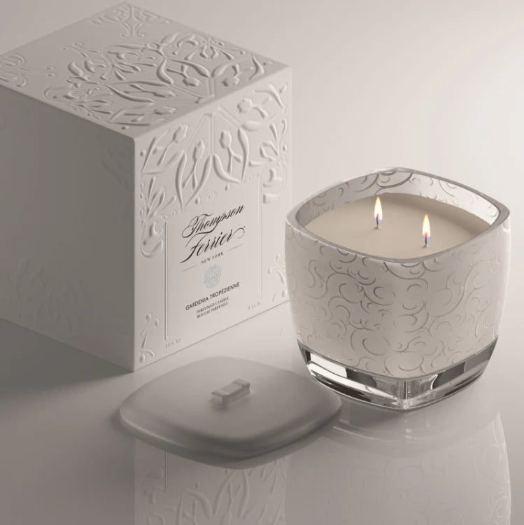 A lit double-wick scented candle in a clear glass jar with a white embossed floral pattern, shown beside its matching white box and lid on a light neutral background.