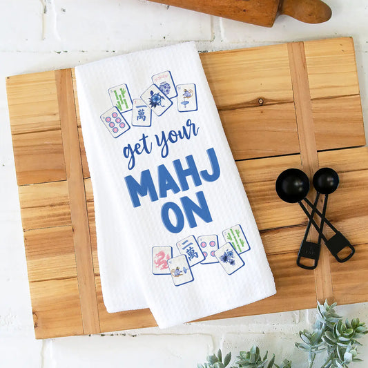 White towel with 'get your MAHJ ON' text and card designs on a wooden cutting board.