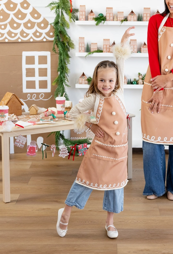 A young girl wearing a tan apron with white trim and buttons stands in a festive gingerbread-themed room, smiling and posing. An adult woman in a matching apron is partially visible on the right.