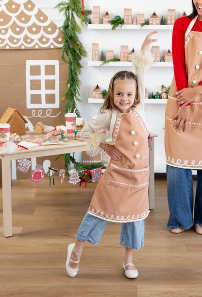 A young girl wearing a tan apron with white trim and buttons stands in a festive gingerbread-themed room, smiling and posing. An adult woman in a matching apron is partially visible on the right.