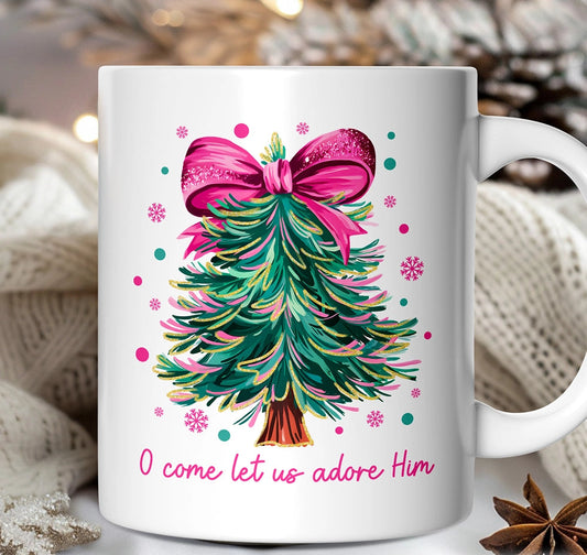 White mug with Christmas tree design and pink bow, featuring text 'O come let us adore Him' on a blurred background.